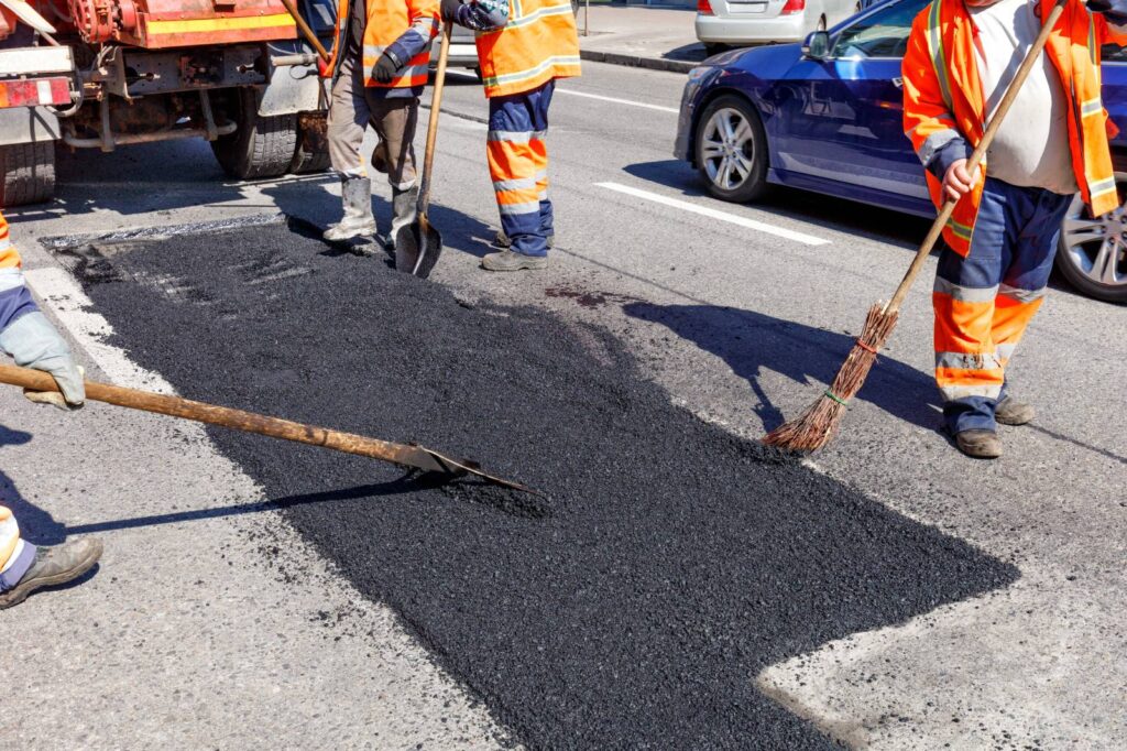 Road maintenance crew repairing a pothole by spreading and leveling fresh hot asphalt on a city street, using shovels and brooms while traffic passes nearby.