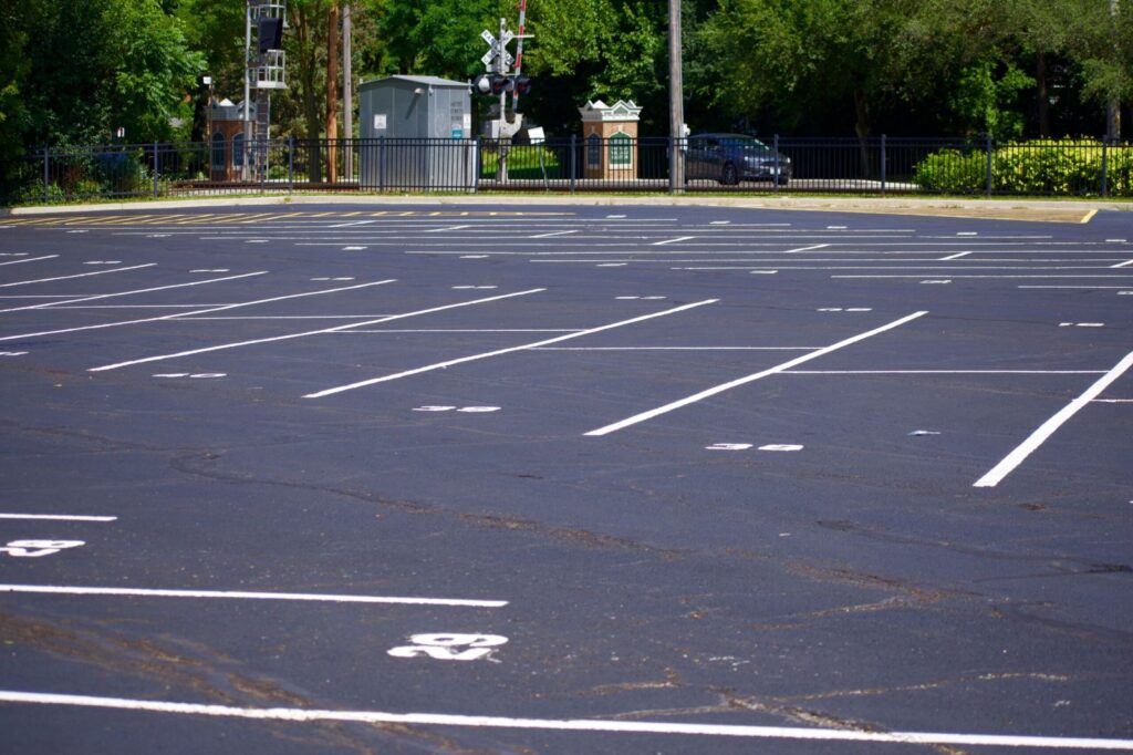 Freshly sealcoated asphalt parking lot with crisp white line striping and numbered parking spaces, set against a backdrop of trees, fencing, and railway crossing signals.