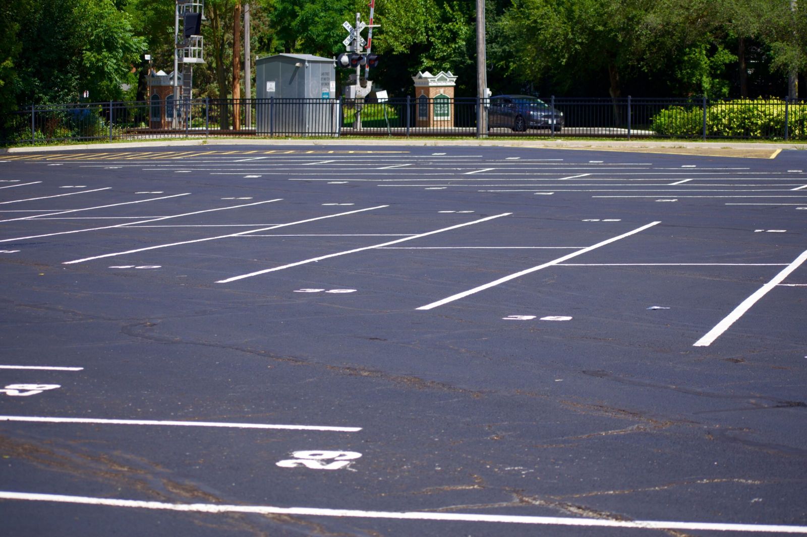 Freshly sealcoated asphalt parking lot with crisp white line striping and numbered parking spaces, set against a backdrop of trees, fencing, and railway crossing signals.
