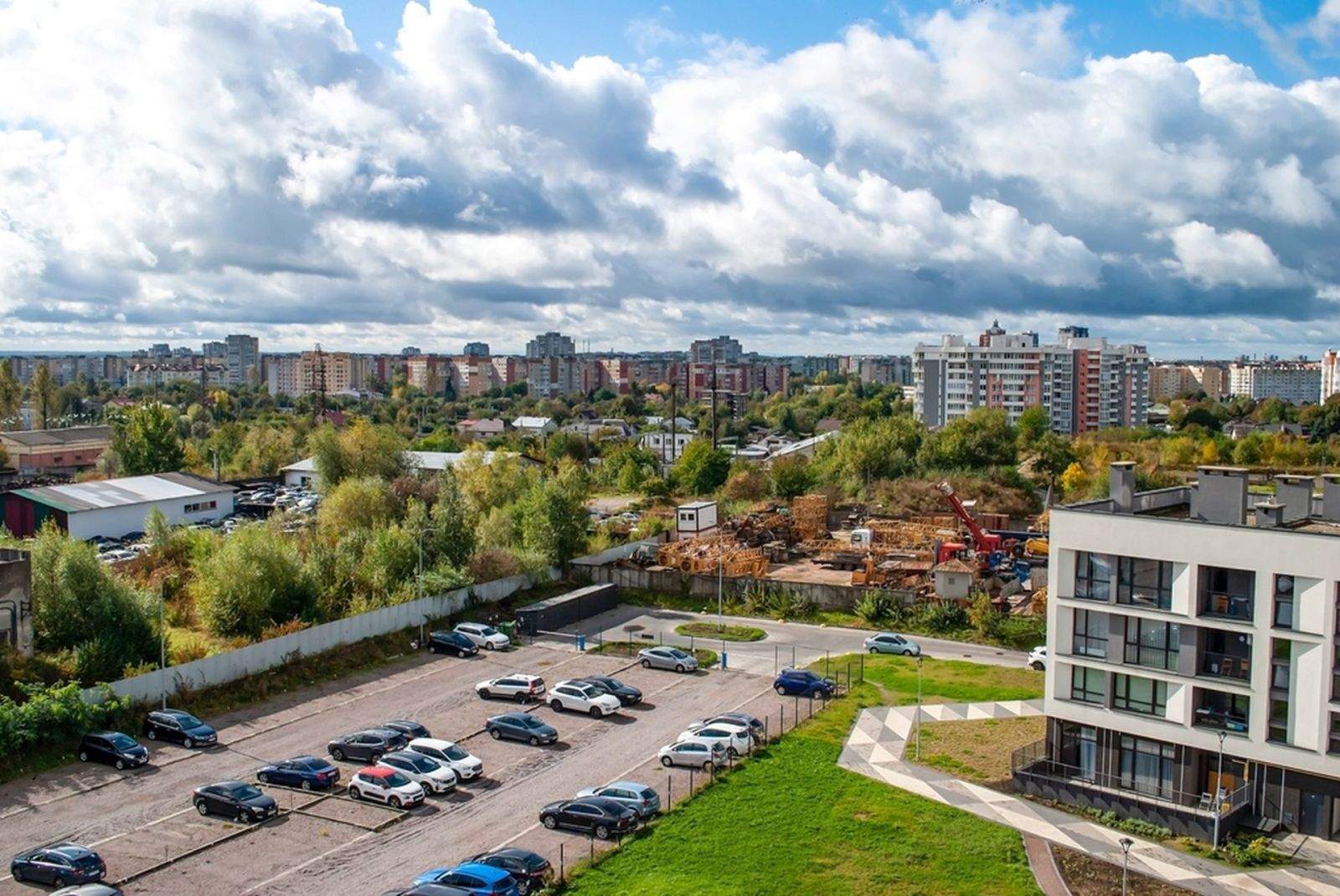 Condo parking lot with multiple parked cars beside a modern residential building, overlooking a mixed urban area with greenery and mid-rise apartments in the background.