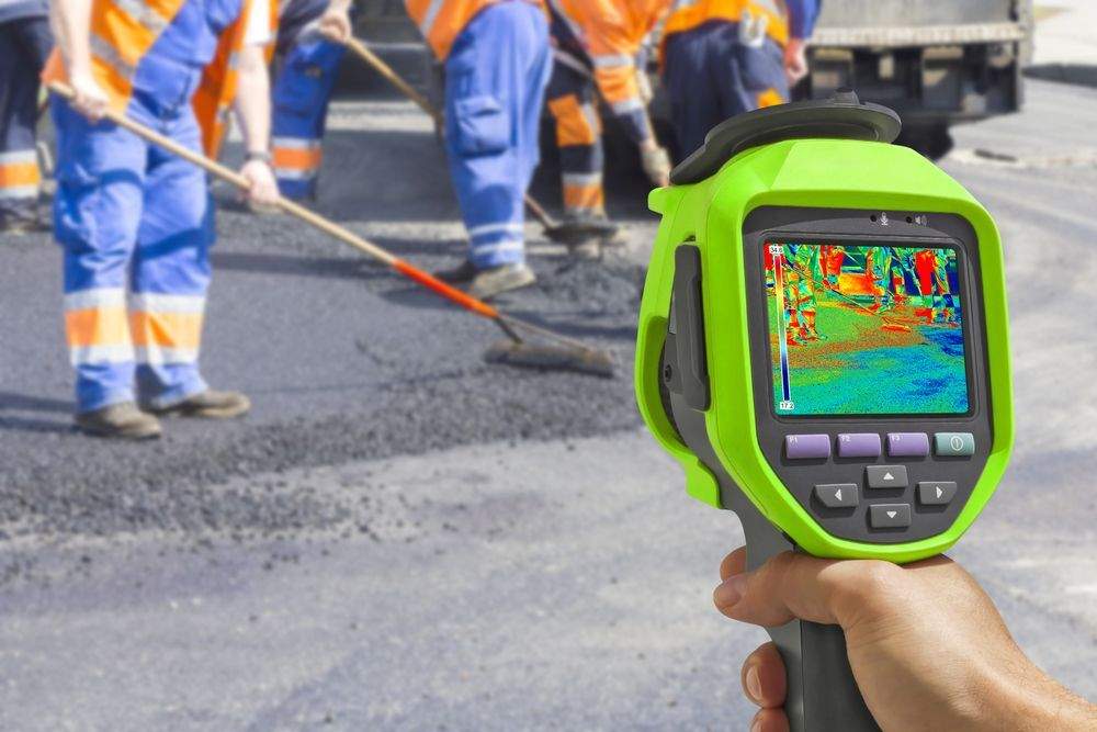 Infrared temperature scanner measuring hot asphalt during industrial pavement repairs in summer, while a paving crew works in the background, highlighting how heat-related asphalt deterioration can be identified and repaired using infrared asphalt restoration techniques.
