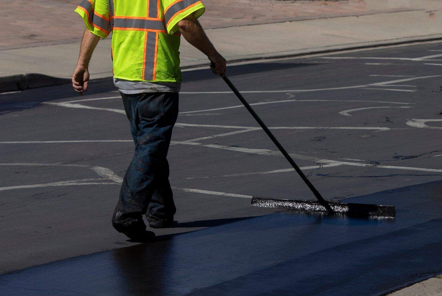 A contractor in a high-visibility safety vest using a large squeegee to apply black asphalt sealcoat over a cracked pavement surface.