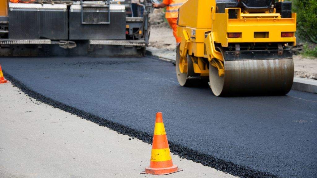 Road roller machine smoothing fresh asphalt alongside a safety cone during a seasonal paving project.